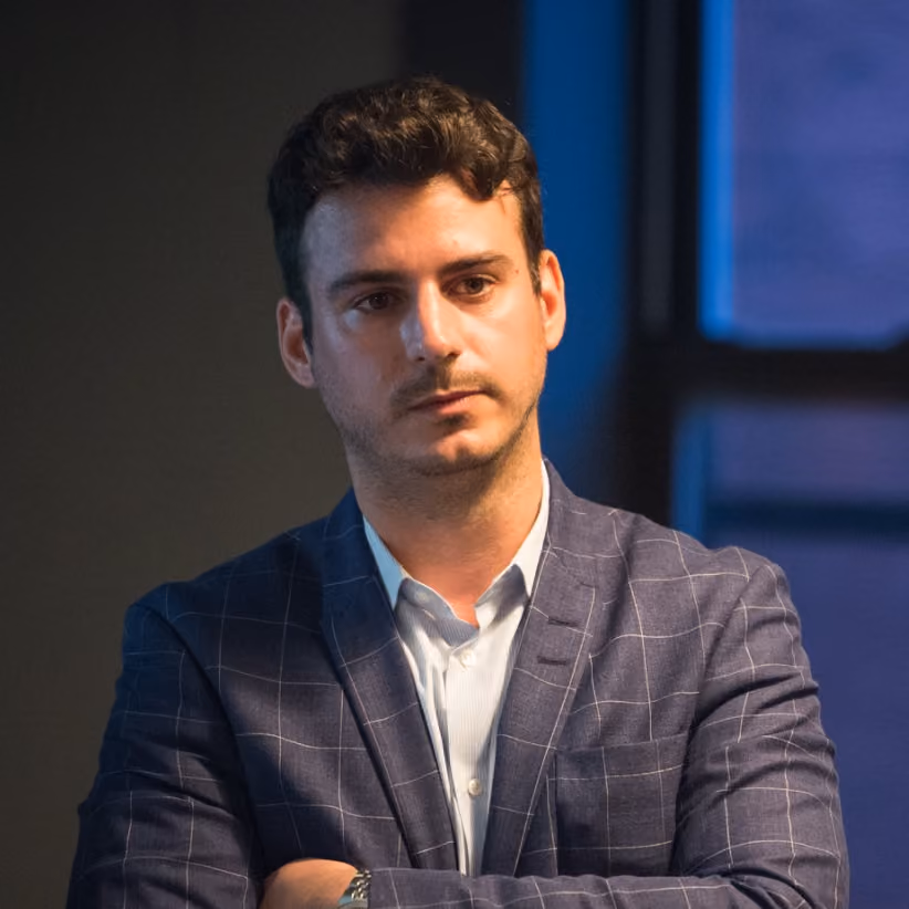 Man with short dark hair and light beard wearing a blue checkered blazer and light shirt standing with arms crossed against a dimly lit background with a window.