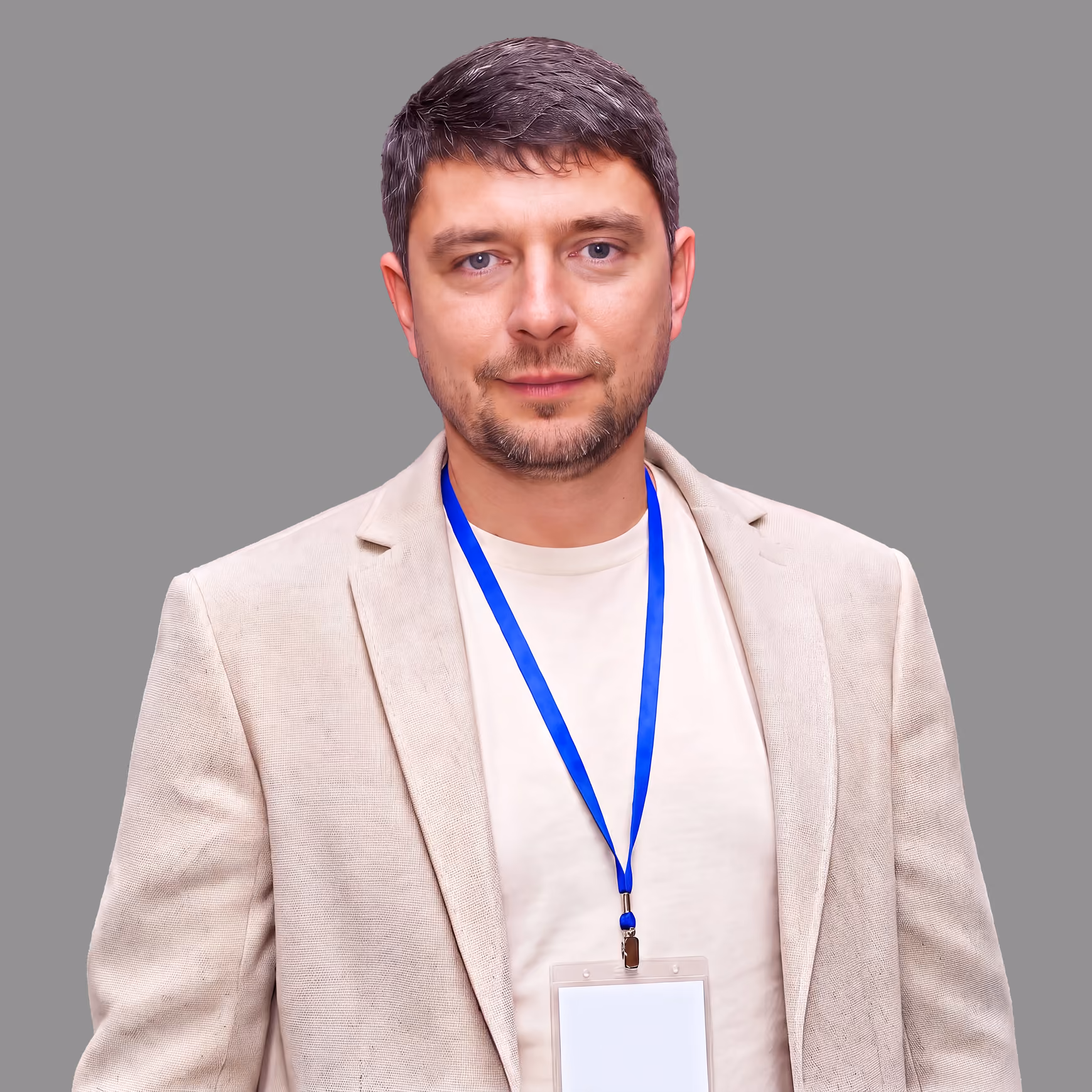 Man with short dark hair and beard wearing a beige blazer and white shirt, with a blue lanyard and blank name badge, against a plain gray background.