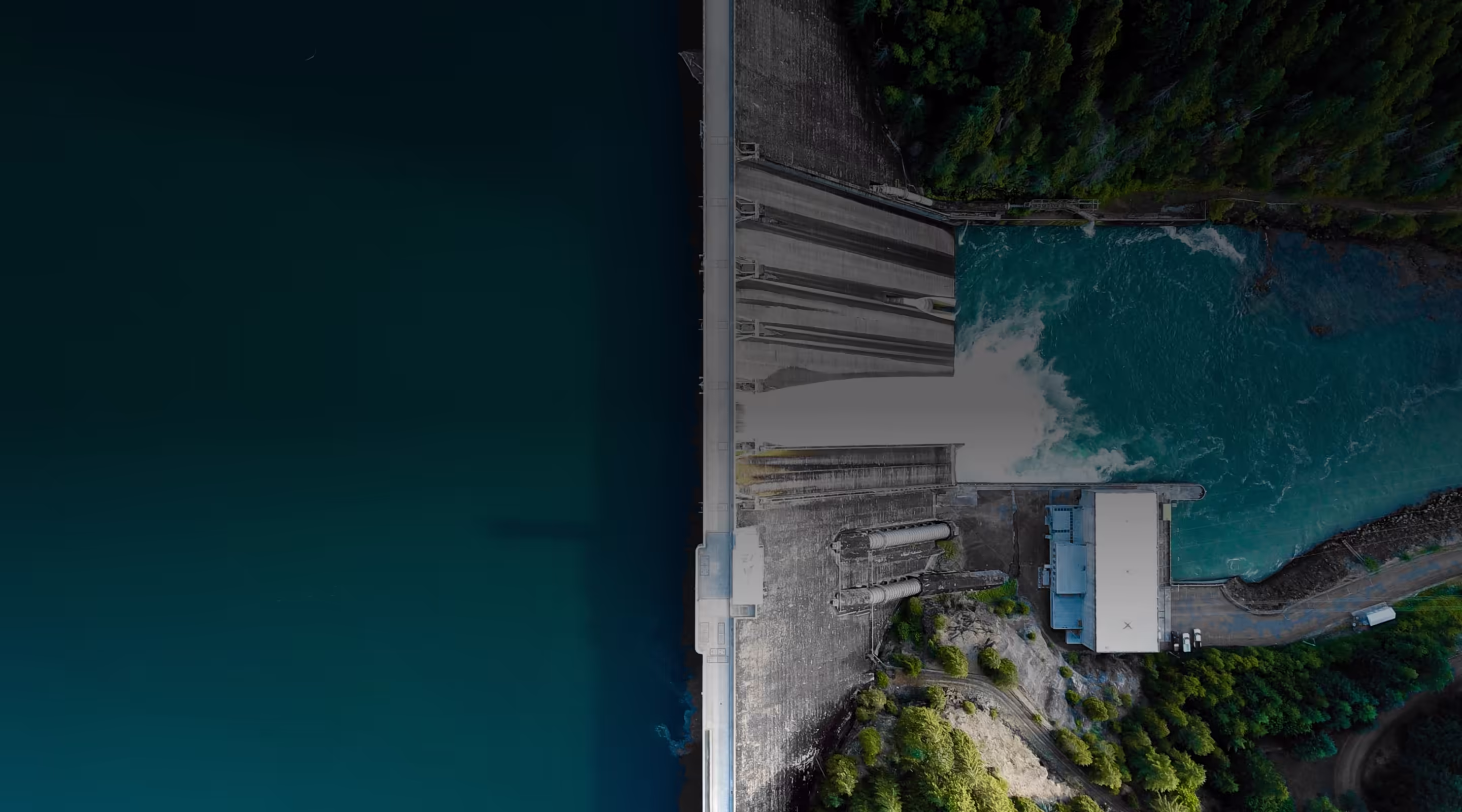Aerial view of a dam releasing water into a turquoise river surrounded by green forest.
