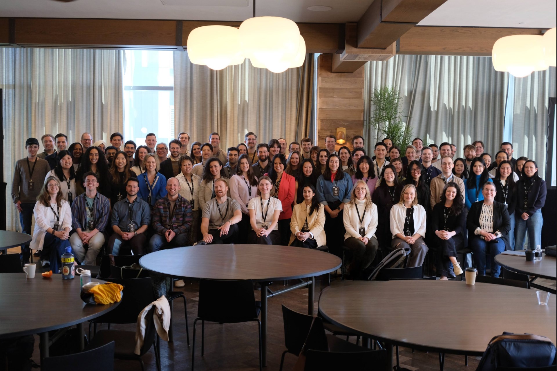 Large diverse group of people posing for a photo in a modern room with round tables and large windows.