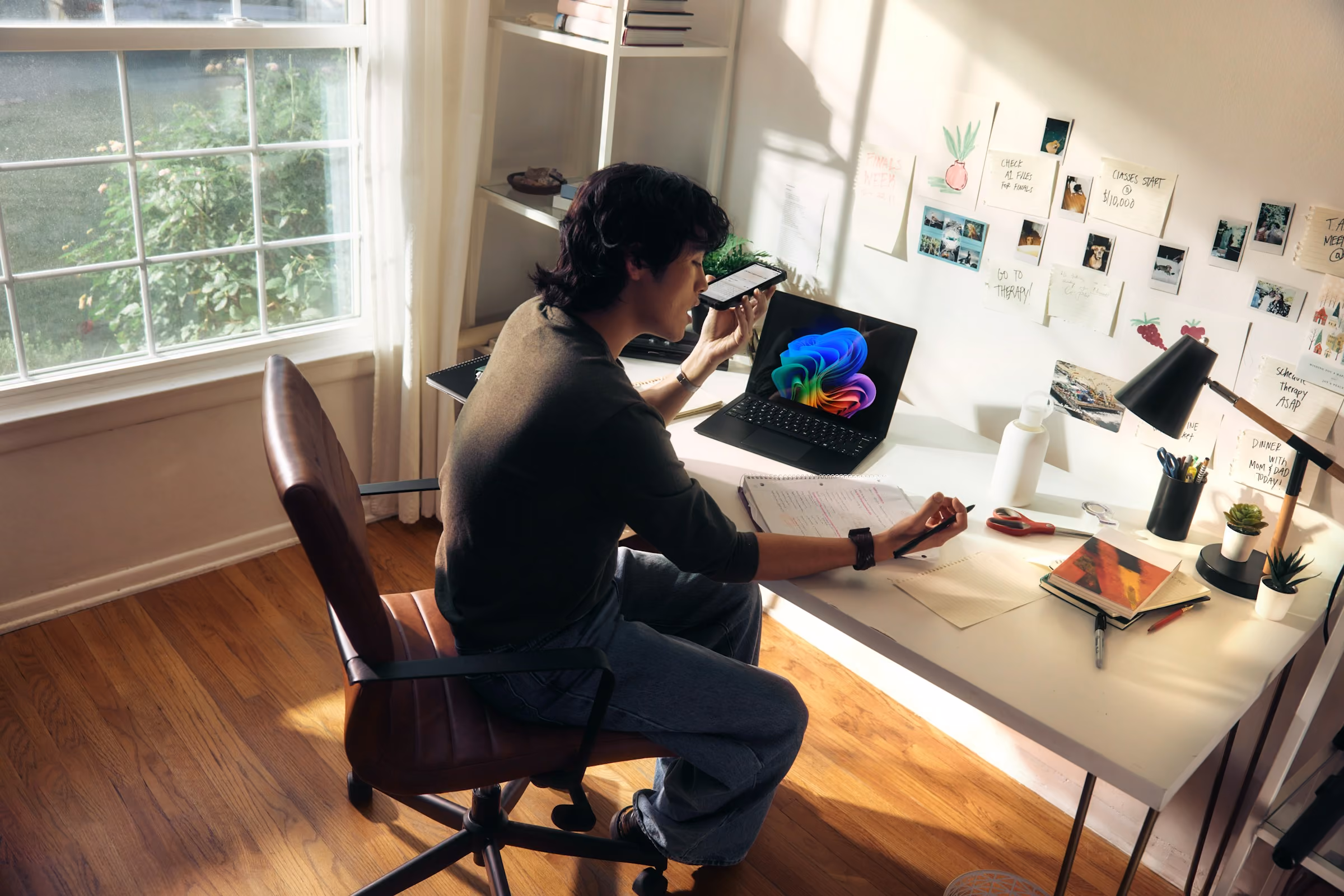 Person sitting at a desk with laptop and notes, speaking into a phone in a sunlit room.