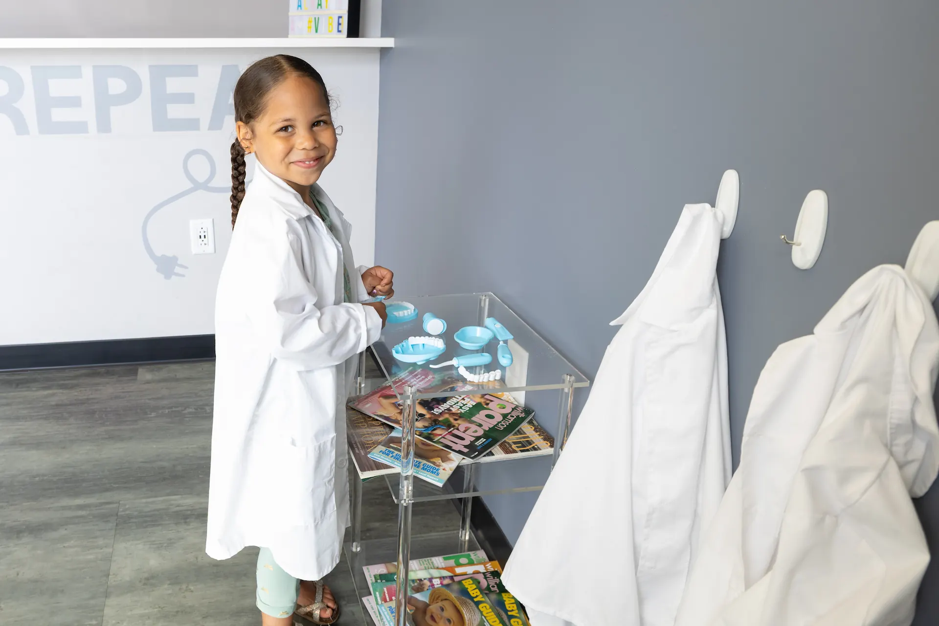 A smiling young girl wearing a white lab coat stands by a clear acrylic table with dental models and magazines, against a gray wall with more white coats hanging on hooks nearby.