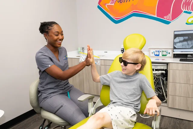 Smiling female dental professional giving a high-five to a young boy sitting in a yellow dental chair in a dental office.