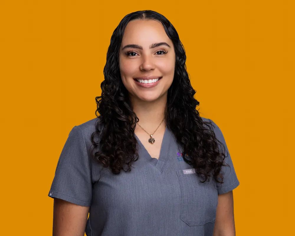 Smiling woman with long curly dark hair wearing gray medical scrubs and a necklace, standing against an orange background.