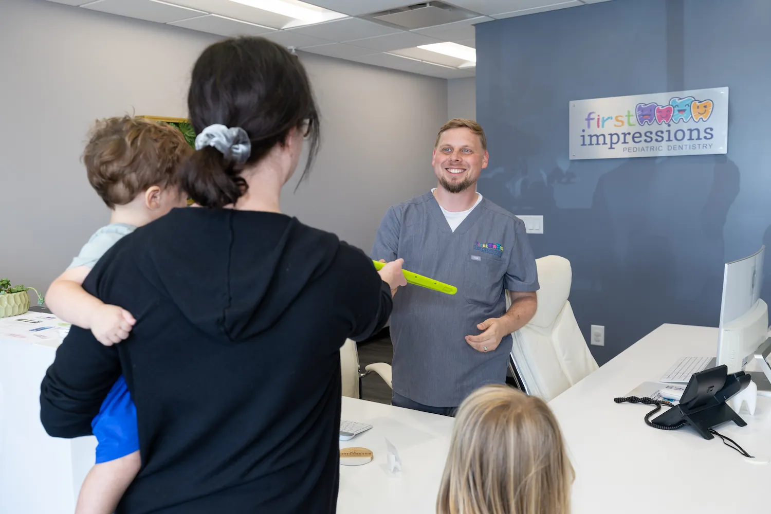 A smiling male pediatric dentist in gray scrubs stands behind a white reception desk while a woman holding a child hands him a green object, with a young girl standing nearby in a dental office labeled First Impressions Pediatric Dentistry.
