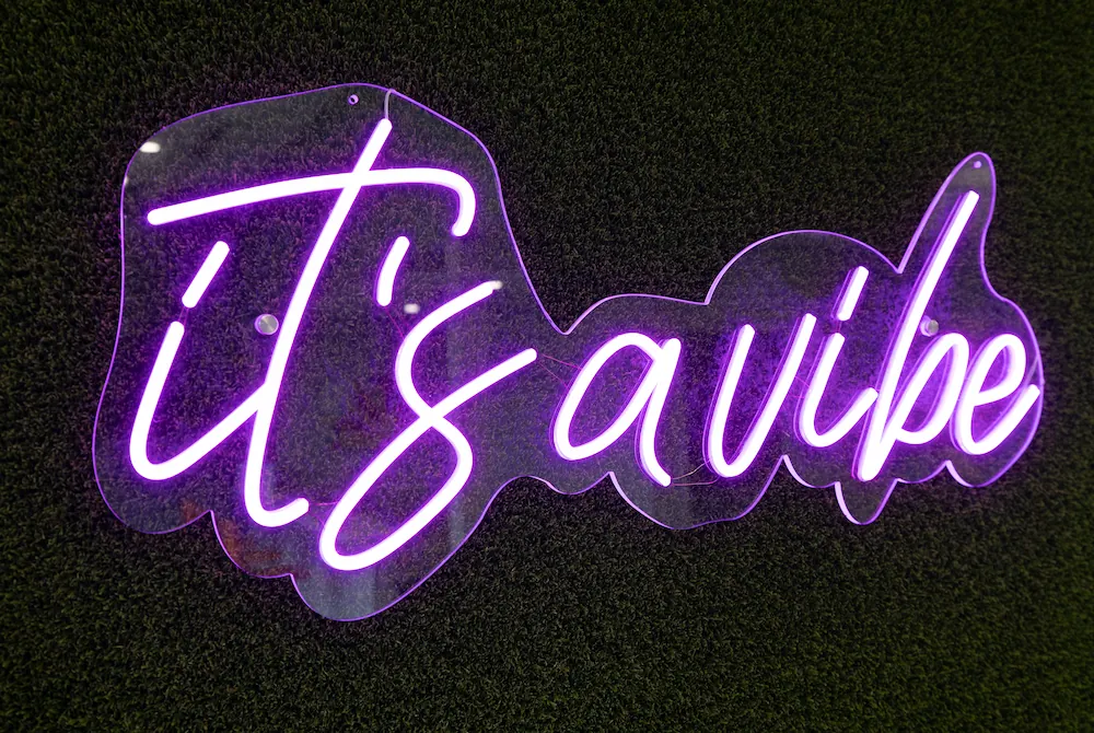 Purple neon sign on a dark textured wall spelling out "it's a vibe" in cursive handwriting.