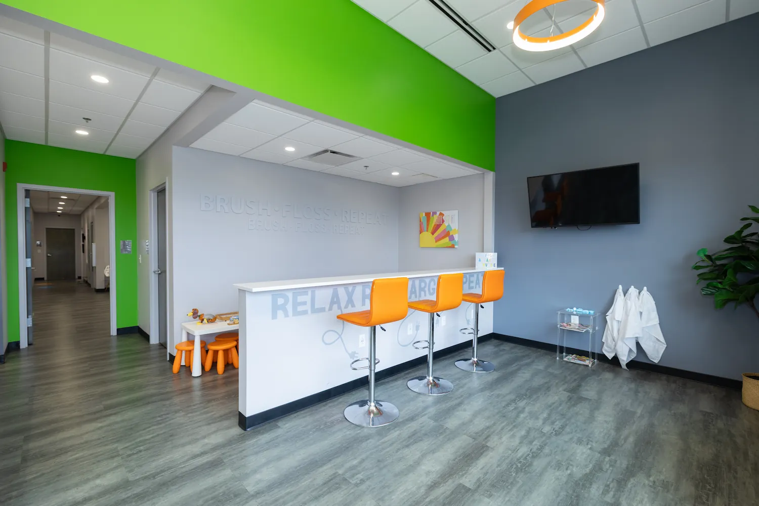 Bright waiting area with three orange bar stools at a white counter that has the words RELAX RECHARGE REPEAT on the front, a TV mounted on a gray wall, a small clear stand holding magazines and items, three white coats hanging below the TV, green accent wall on the left, children's play table with orange stools in a corner, and a ceiling light fixture above the counter.