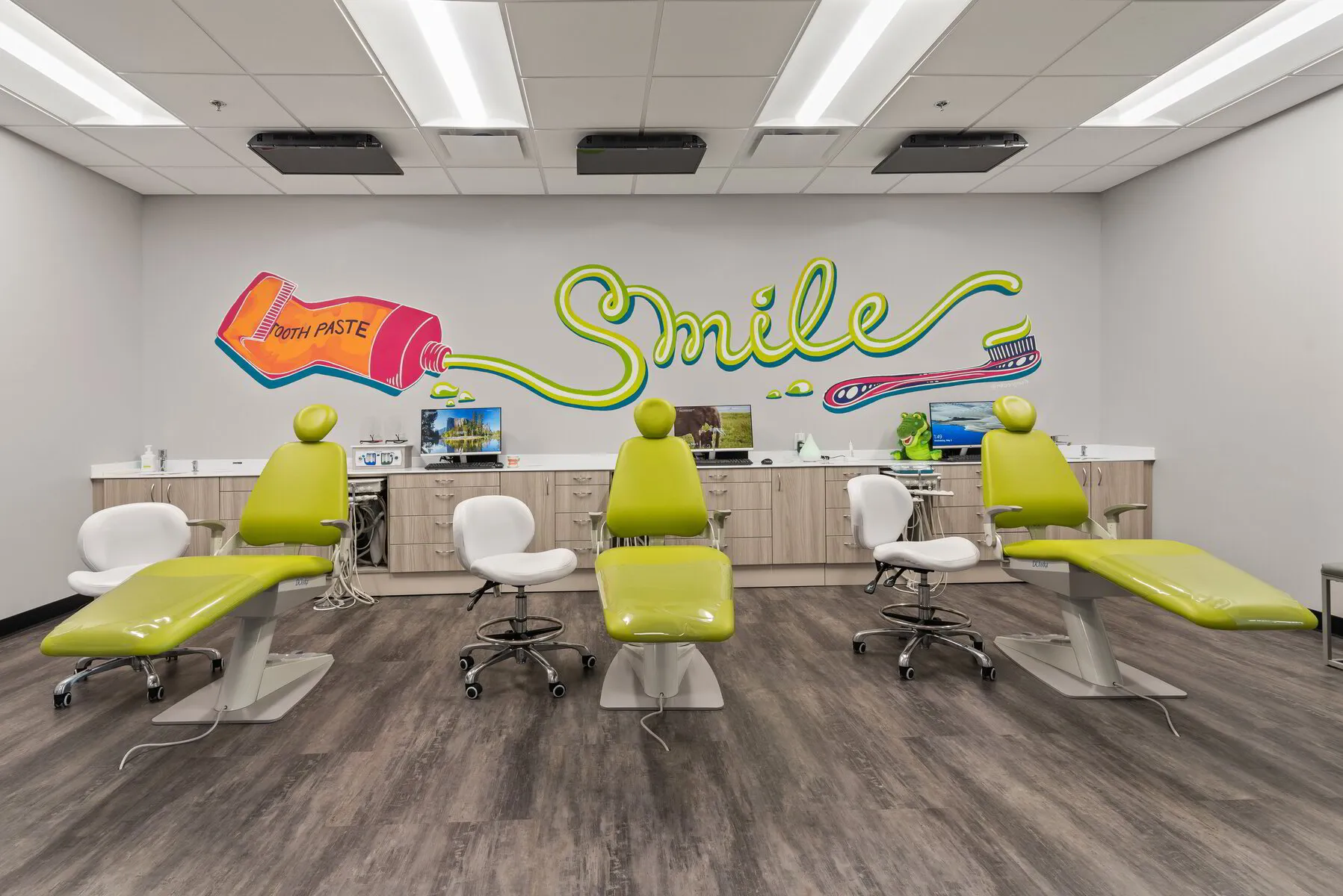 Modern dental office with three lime green dental chairs and white stools against a wall featuring a colorful mural of a toothpaste tube, toothbrush, and the word 'Smile' in vibrant lettering.