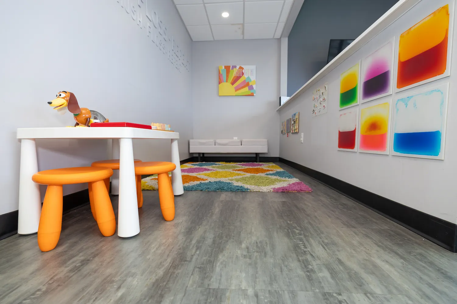 Children's playroom with a small white table and two orange stools, a Shaggy dog toy on the table, colorful geometric carpet, white bench against gray wall, and vibrant abstract artwork on the side wall.