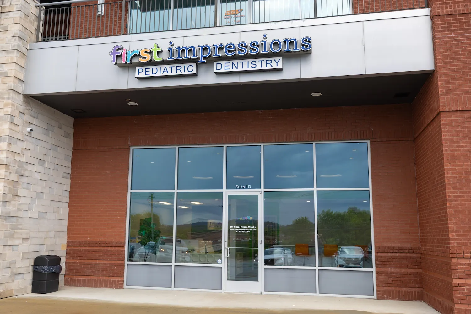 Front entrance of First Impressions Pediatric Dentistry showing a glass door and windows framed by red brick, with colorful lettering of the word 'first' and blue 'impressions', and the words 'PEDIATRIC DENTISTRY' in blue on white panels above the door.