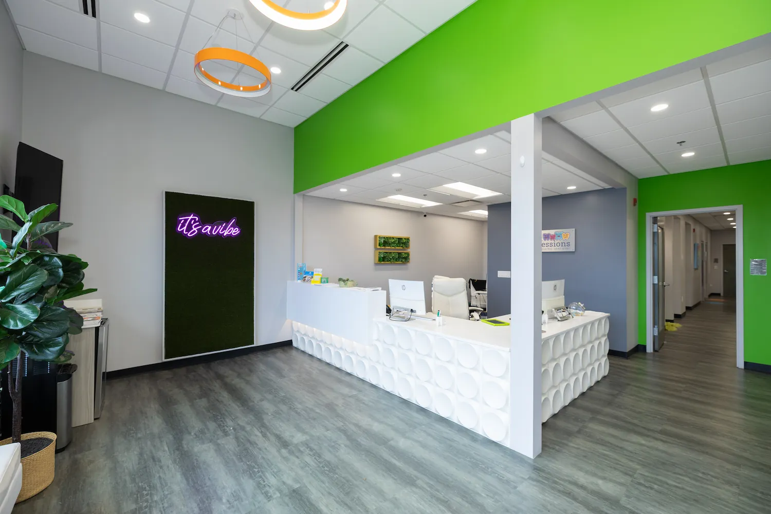 Modern office reception area with a white desk featuring circular patterns, green accent wall, potted plant, and a neon sign reading 'it's a vibe' on a black background.