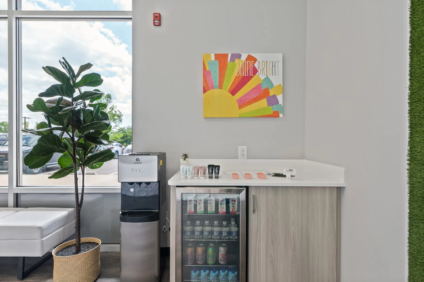 A modern refreshment corner with a large window showing cars outside, a tall green plant in a woven basket, an Avalon water cooler, a mini fridge stocked with beverages, and a countertop holding various product samples and soap bars, with a colorful 'Shine Bright' wall art above.