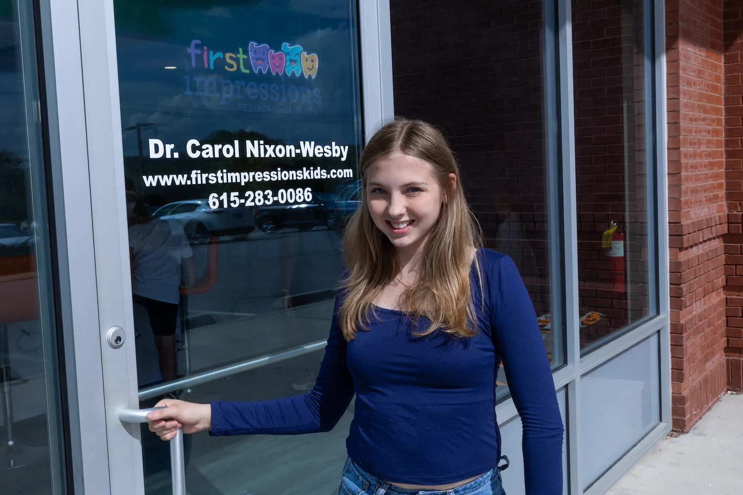Young woman with long blonde hair wearing a navy blue long-sleeve top and jeans, standing outside and holding the door handle of a glass door featuring a sign for Dr. Carol Nixon-Wesby, a pediatric dentist at First Impressions with the website and phone number displayed.
