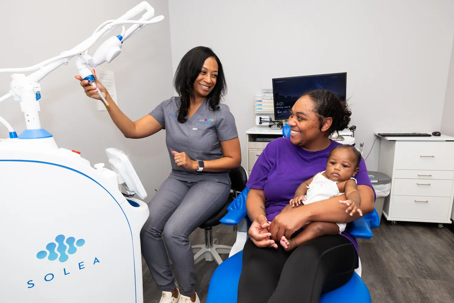 A female dental professional in gray scrubs adjusts a dental laser machine while a woman in a purple shirt holds a baby sitting on a blue dental chair, all smiling inside a modern dental office.