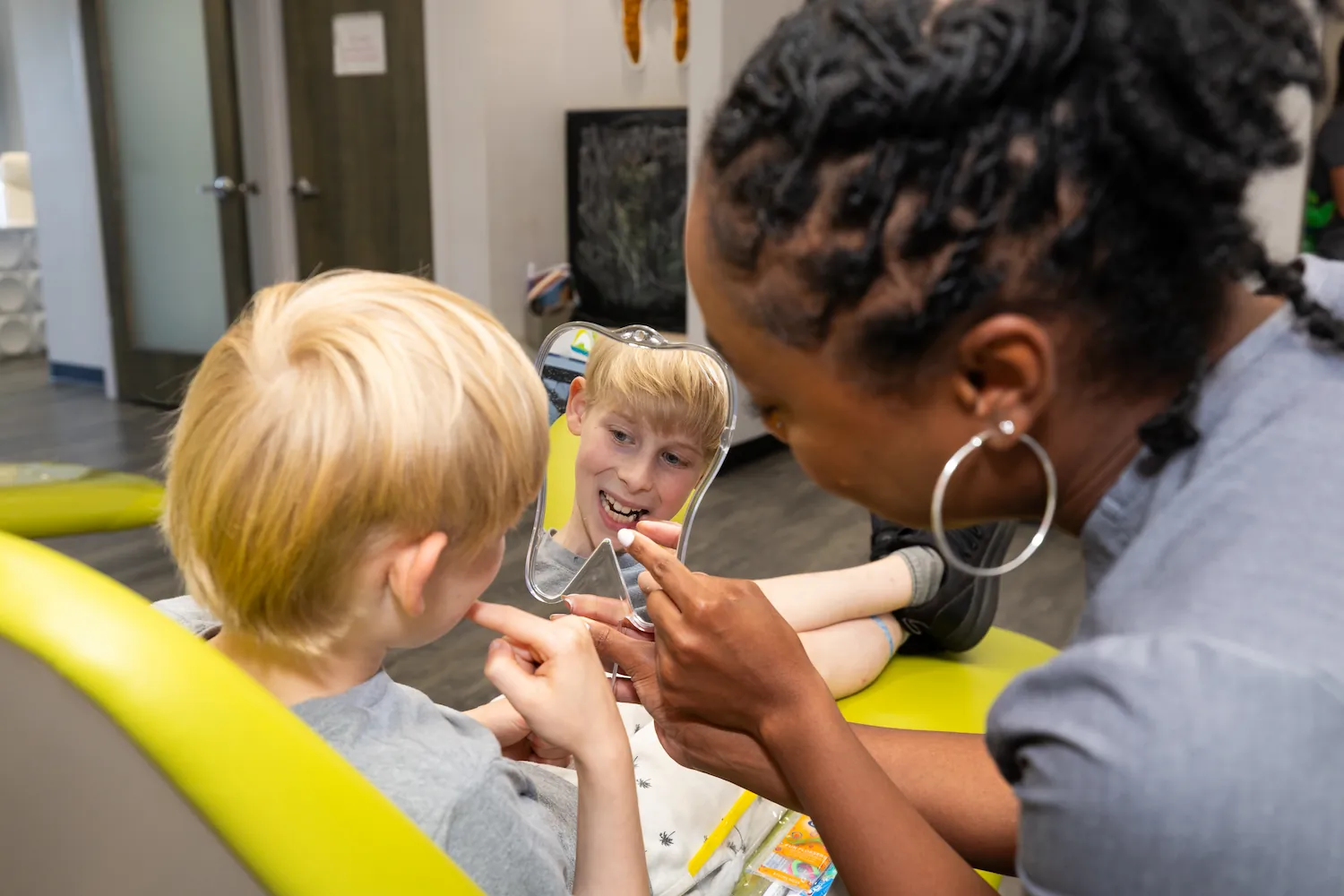 A smiling blonde boy sitting in a yellow dental chair looking into a tooth-shaped mirror held by a woman with braided hair and large hoop earrings showing him a dental item.