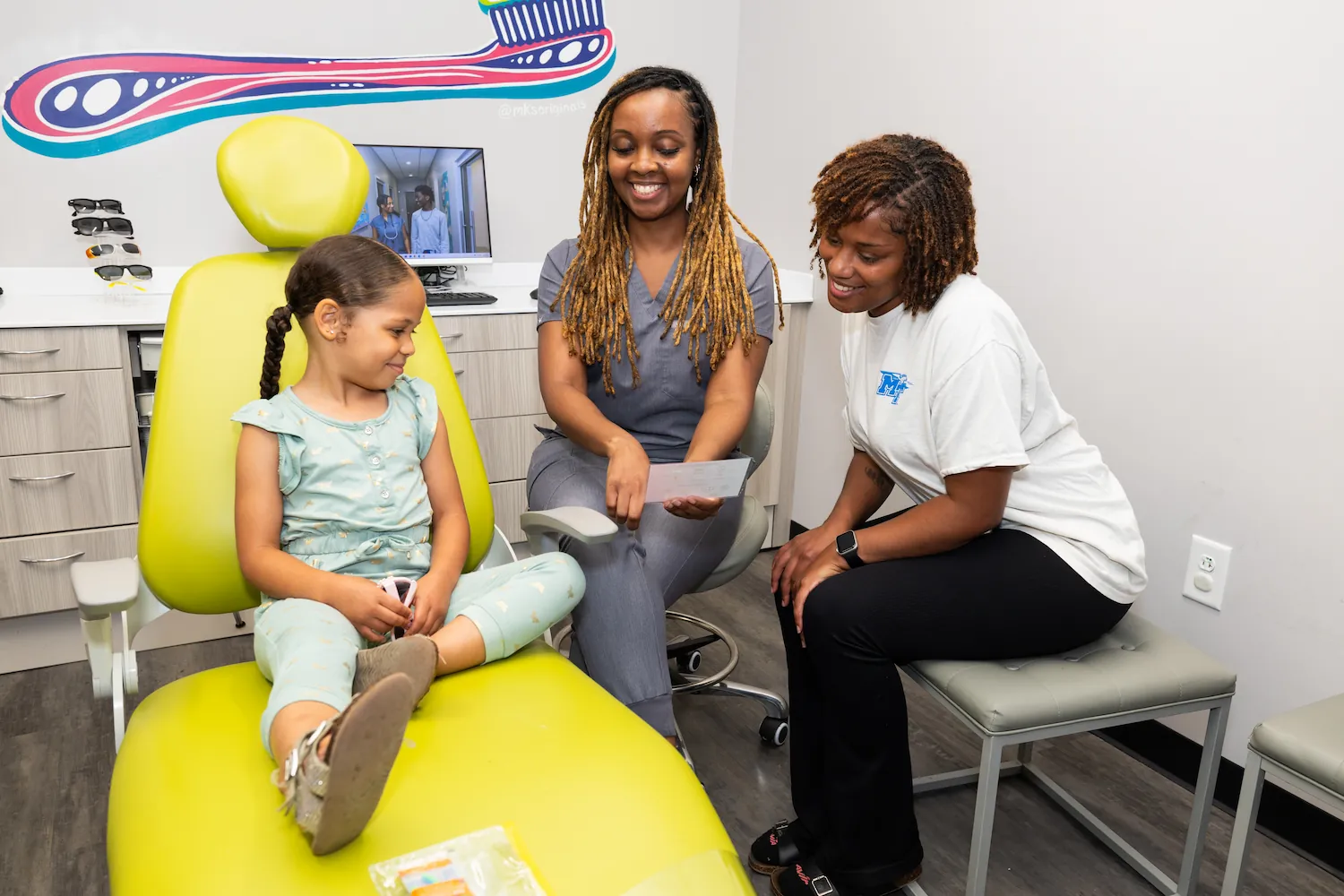 A young girl sits on a bright green dental chair while a female dental professional in gray scrubs and another woman in a white shirt talk and smile with her in a dental office. A large colorful toothbrush wall decoration is visible above a computer monitor in the background.