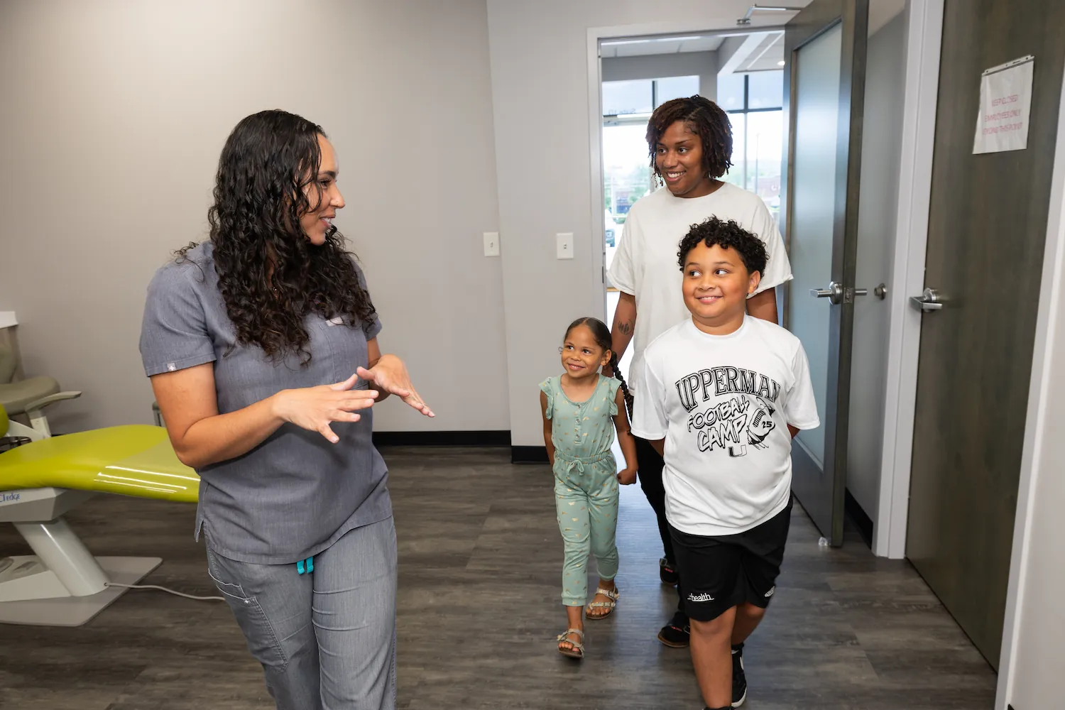 A female healthcare worker in gray scrubs speaks and gestures with her hands to a woman and two children walking through a medical office hallway; the woman and children smile and listen attentively.