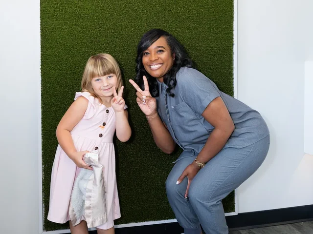 A smiling woman and young girl stand in front of a green textured backdrop, both holding up peace signs; the woman is crouched slightly and wears a gray outfit, while the girl wears a light pink dress and holds a cloth.