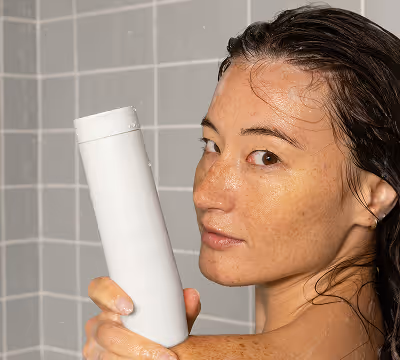 Woman with wet hair holding a white shampoo bottle in a gray tiled shower.