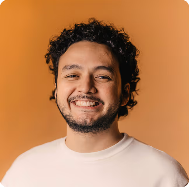 Smiling young man with curly hair and beard wearing a white shirt against an orange background.