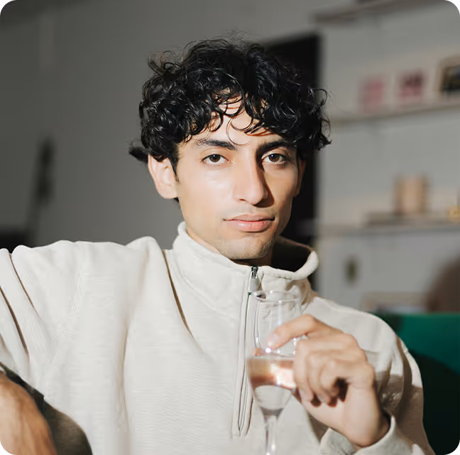 Young man with curly dark hair wearing a light-colored zip-up sweater holding a glass of water indoors.