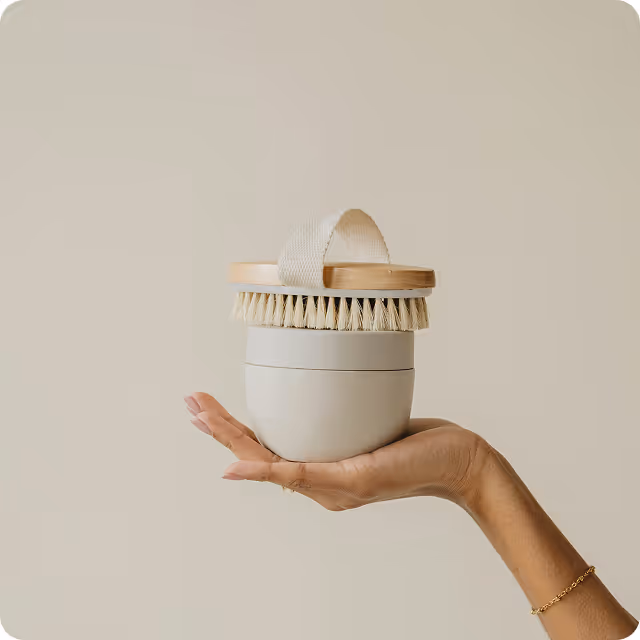 Hand holding a natural bristle body brush with a wooden handle and a white ceramic container against a beige background.