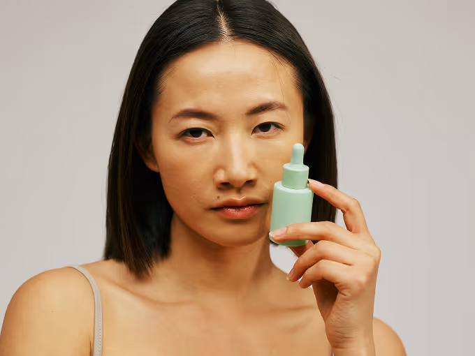 Woman with straight dark hair holding a mint green dropper bottle near her face.