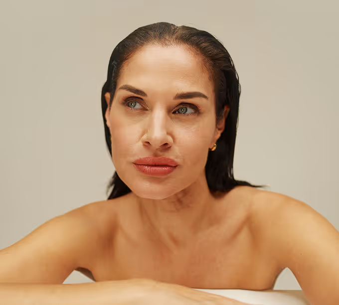 Woman with wet hair, bare shoulders, and gold earrings gazing thoughtfully to the side.