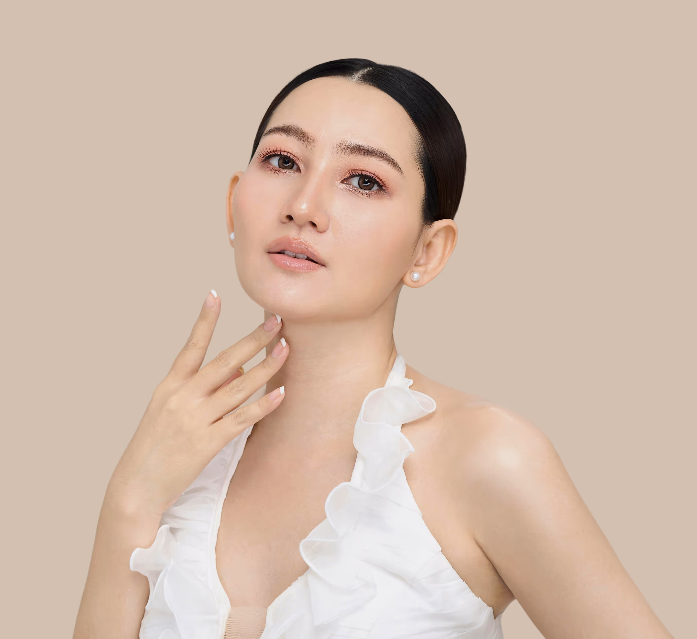 Close-up of a woman with smooth skin, subtle makeup, and pearl earrings wearing a white ruffled halter top against a beige background.