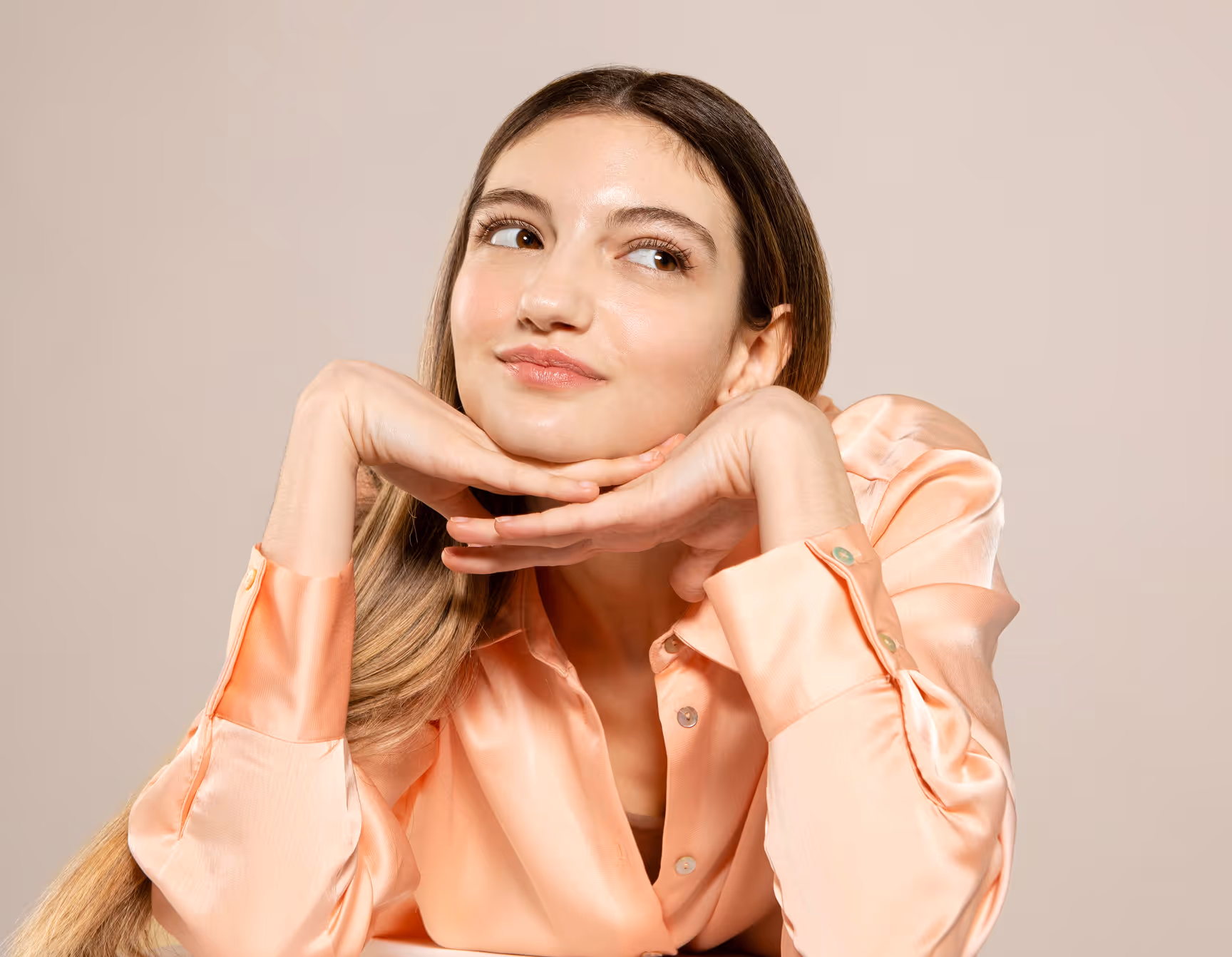 Woman with long hair and peach satin blouse smiling with her chin resting on her hands.