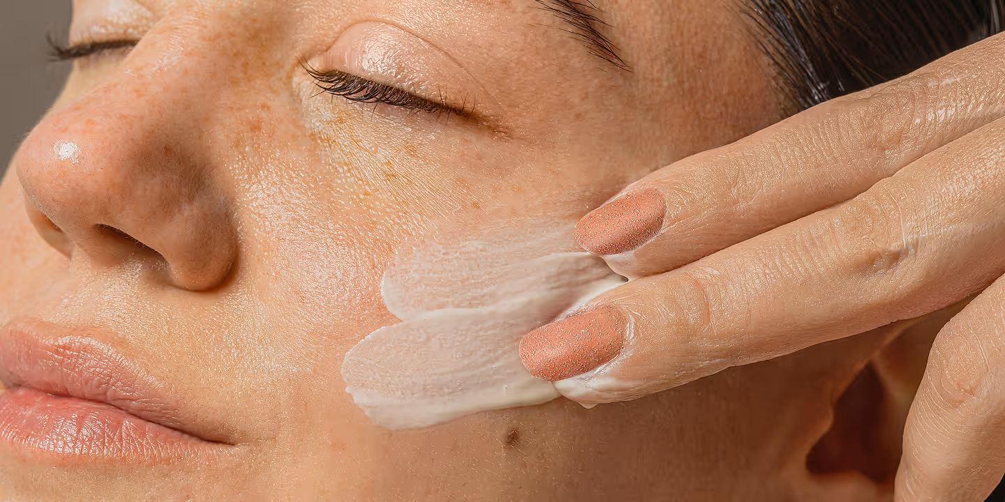 Close-up of a person applying white cream to their cheek with fingers having coral nail polish.
