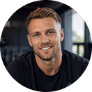 Mann mit Bart und kurzem Haar lächelt in die Kamera, trägt schwarzes T-Shirt, Fitnessstudio-Hintergrund.