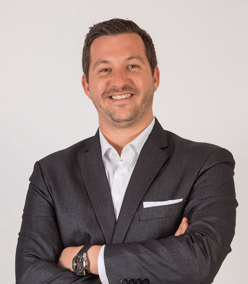 Mann im Anzug lächelt mit verschränkten Armen, weißes Hemd, Armbanduhr, neutraler Hintergrund.