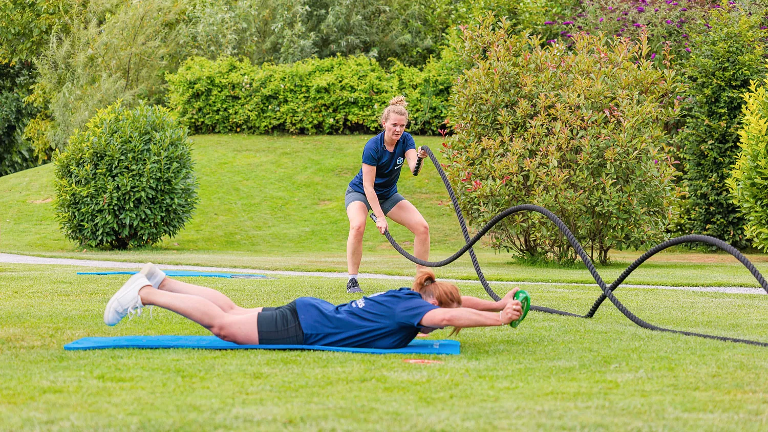 Twee collega's trainen buiten op het gras van de Blue Bricks campus met battle ropes en een buikspierwiel — lage-resolutiebestand (1500 px breed), hoge-resolutieversie nog op te vragen bij de klant
