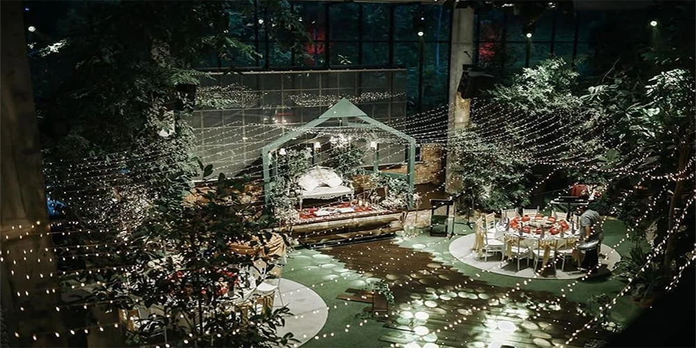 Indoor event space decorated with string lights overhead, round dining tables with chairs, and greenery, featuring a small stage with white cushioned seating inside a green-framed structure.