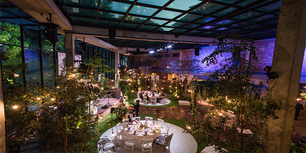 Indoor event space with a glass ceiling and large windows, decorated with trees and string lights; round tables with white chairs are set for dining amid lush greenery and warm ambient lighting.