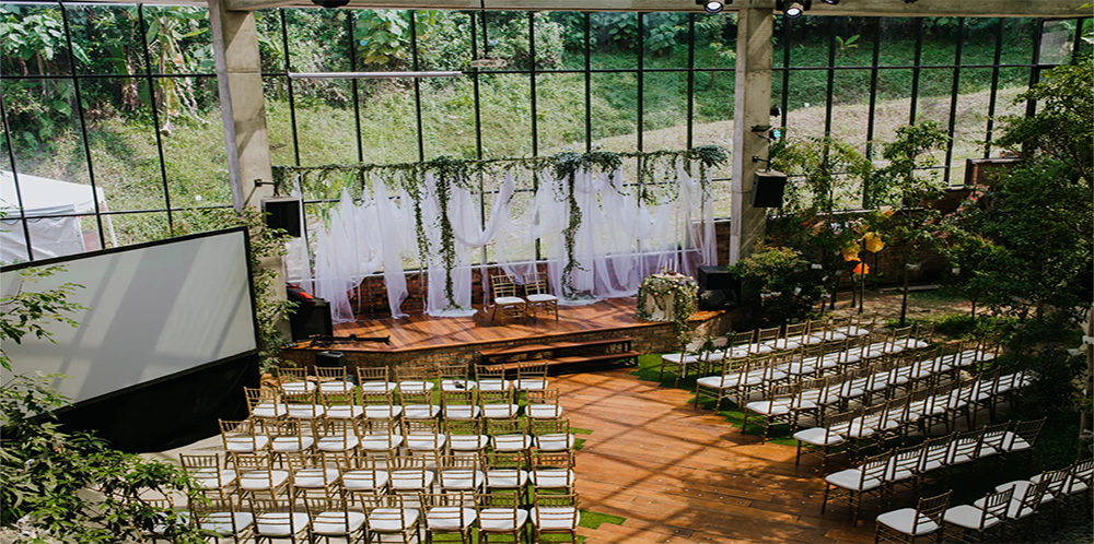 Interior of a glasshouse set up for a wedding ceremony with rows of gold chairs with white cushions arranged facing a small wooden stage decorated with white drapes and greenery.