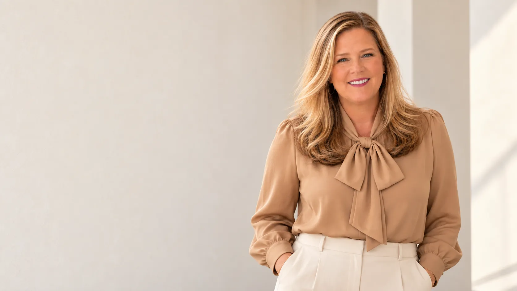 Polly Keene smiling in a tan bow blouse and cream trousers against a soft neutral wall