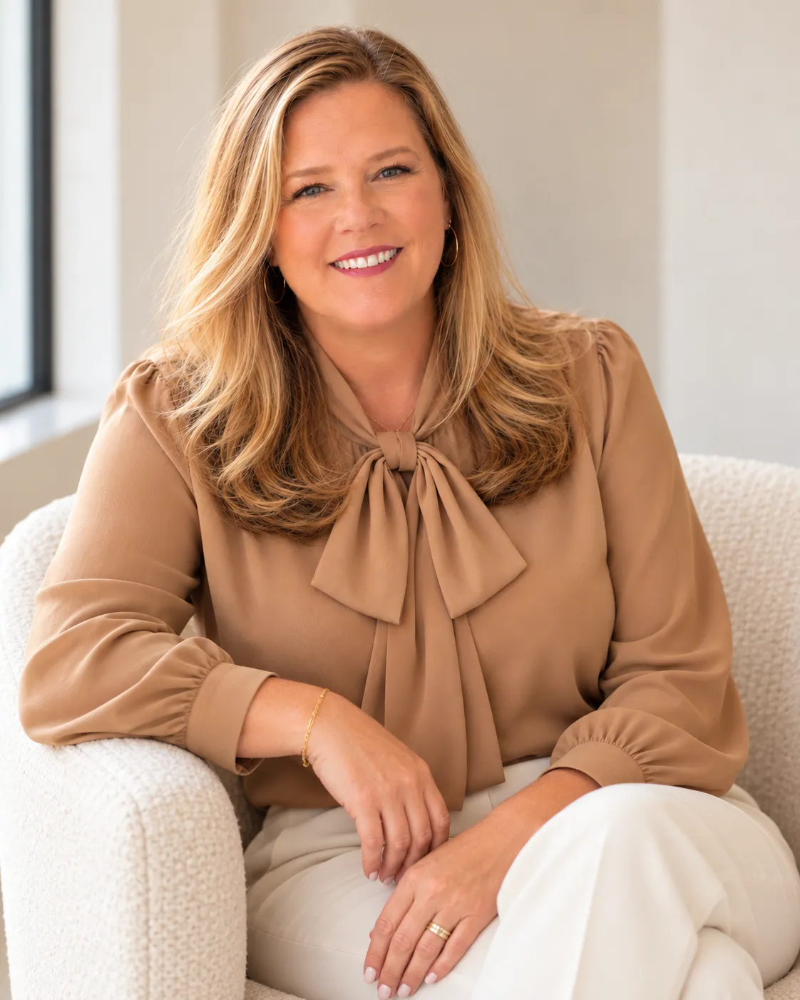 Polly Keene smiling in a tan bow blouse, seated in a cream boucle chair by a window