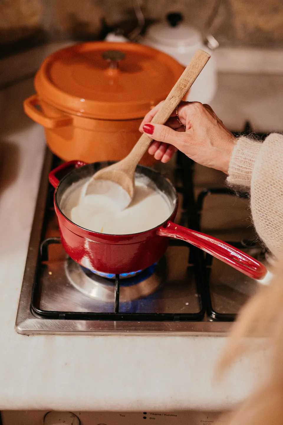 A person stirring a white sauce in a red saucepan on a gas stove