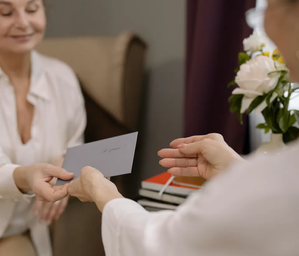 One woman handing a gift envelope to another, who reaches out to receive it, flowers in the background