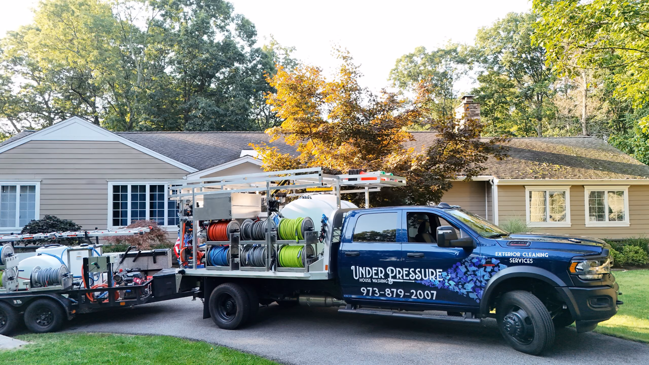 Blue truck with house washing equipment parked in front of a beige house with trees in the background.