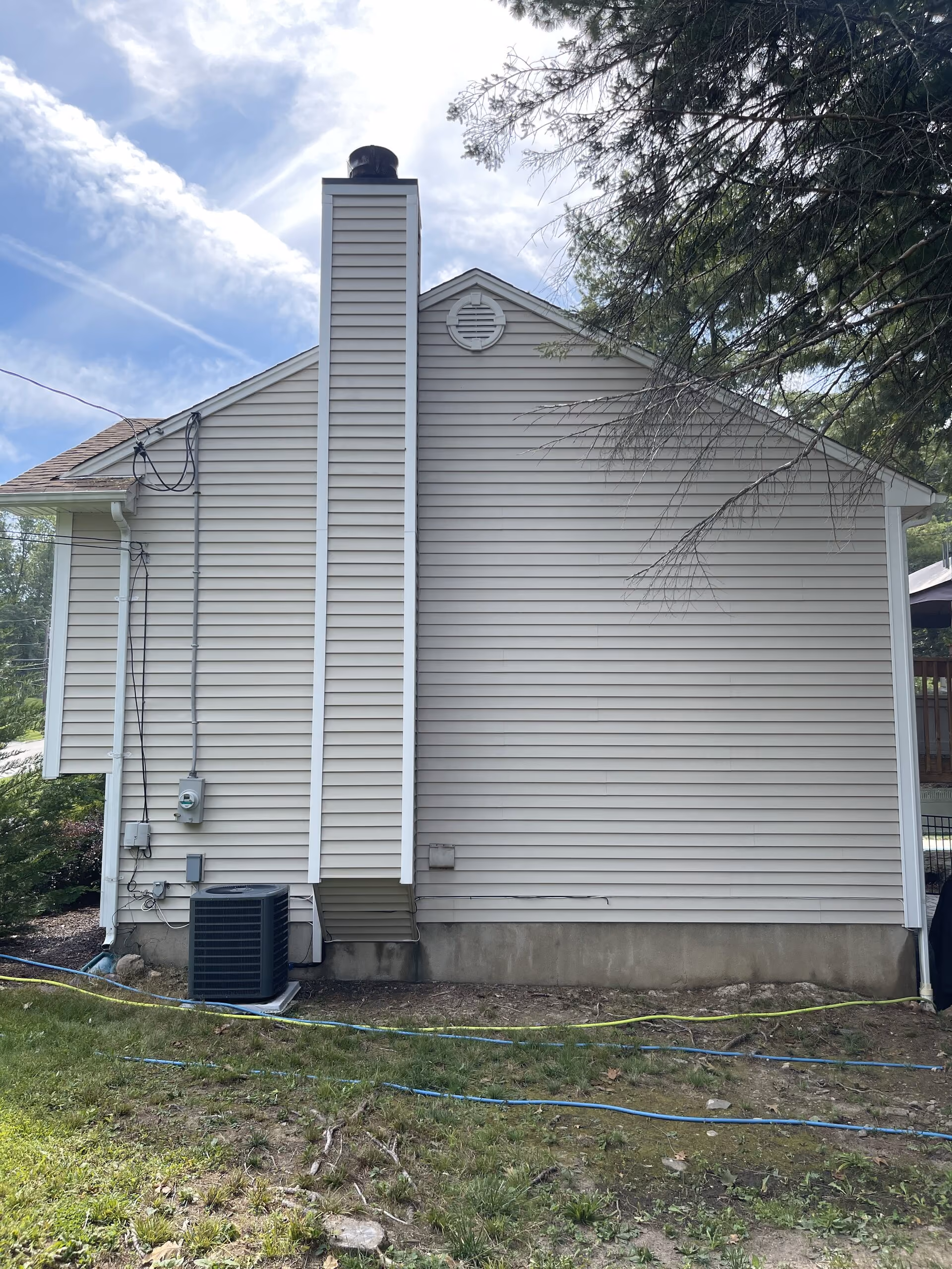 Side of a beige house with chimney, air conditioning unit, and grassy yard under blue sky.