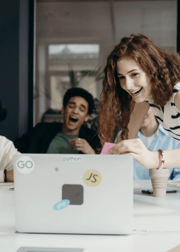 Young woman smiling and leaning over a laptop with programming stickers while others laugh in background.