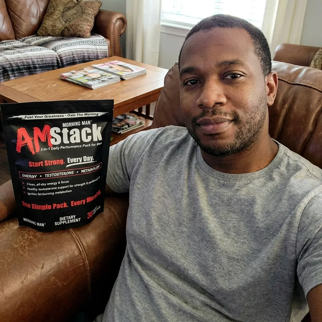 Man in gray t-shirt sitting on brown leather armchair holding a black package of AM Stack dietary supplement for men, with a wooden coffee table and couch in the background.