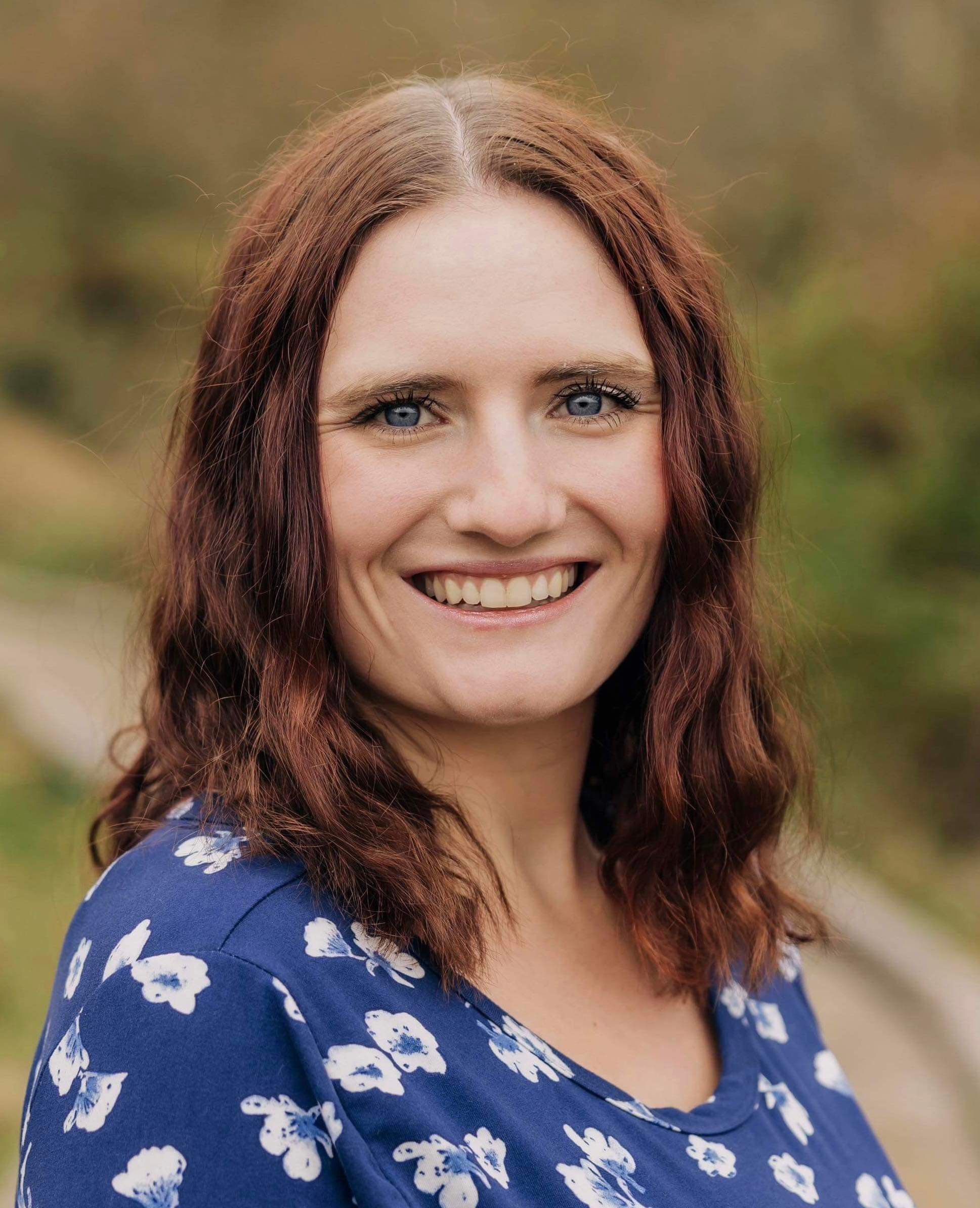 Smiling woman with wavy brown hair and blue eyes wearing a blue top with white flowers outdoors.