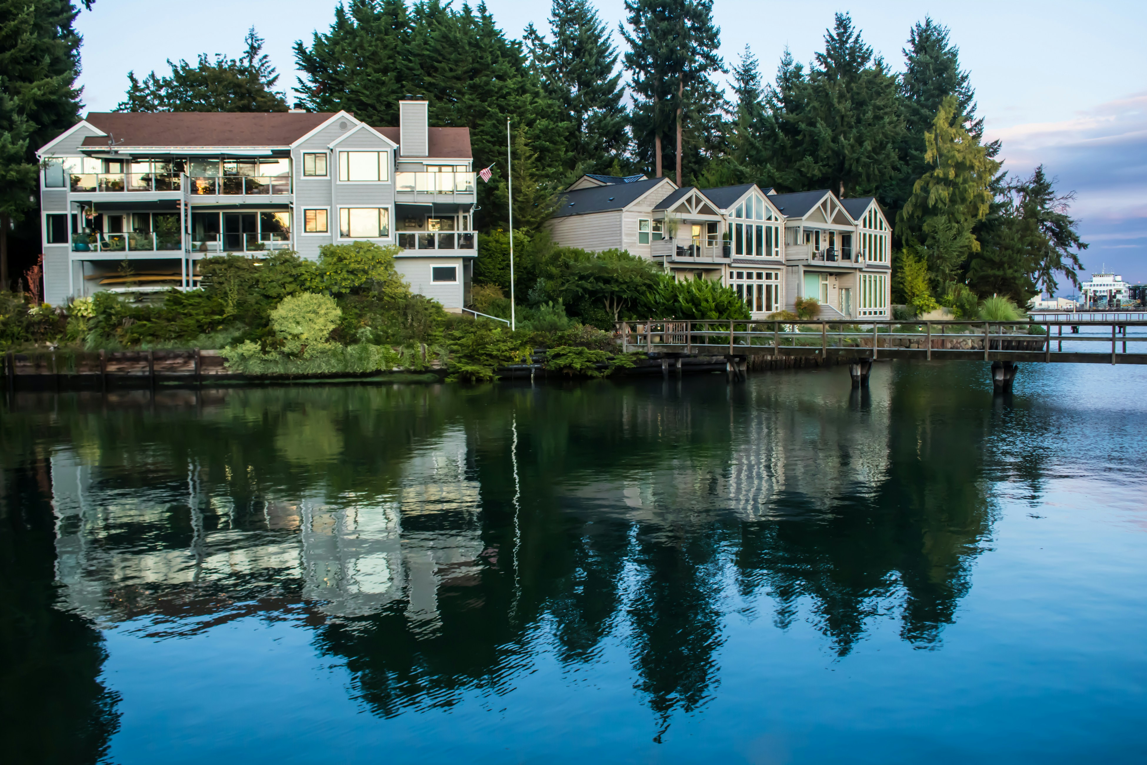 Two waterfront houses with large windows reflect on calm water, surrounded by trees and greenery.