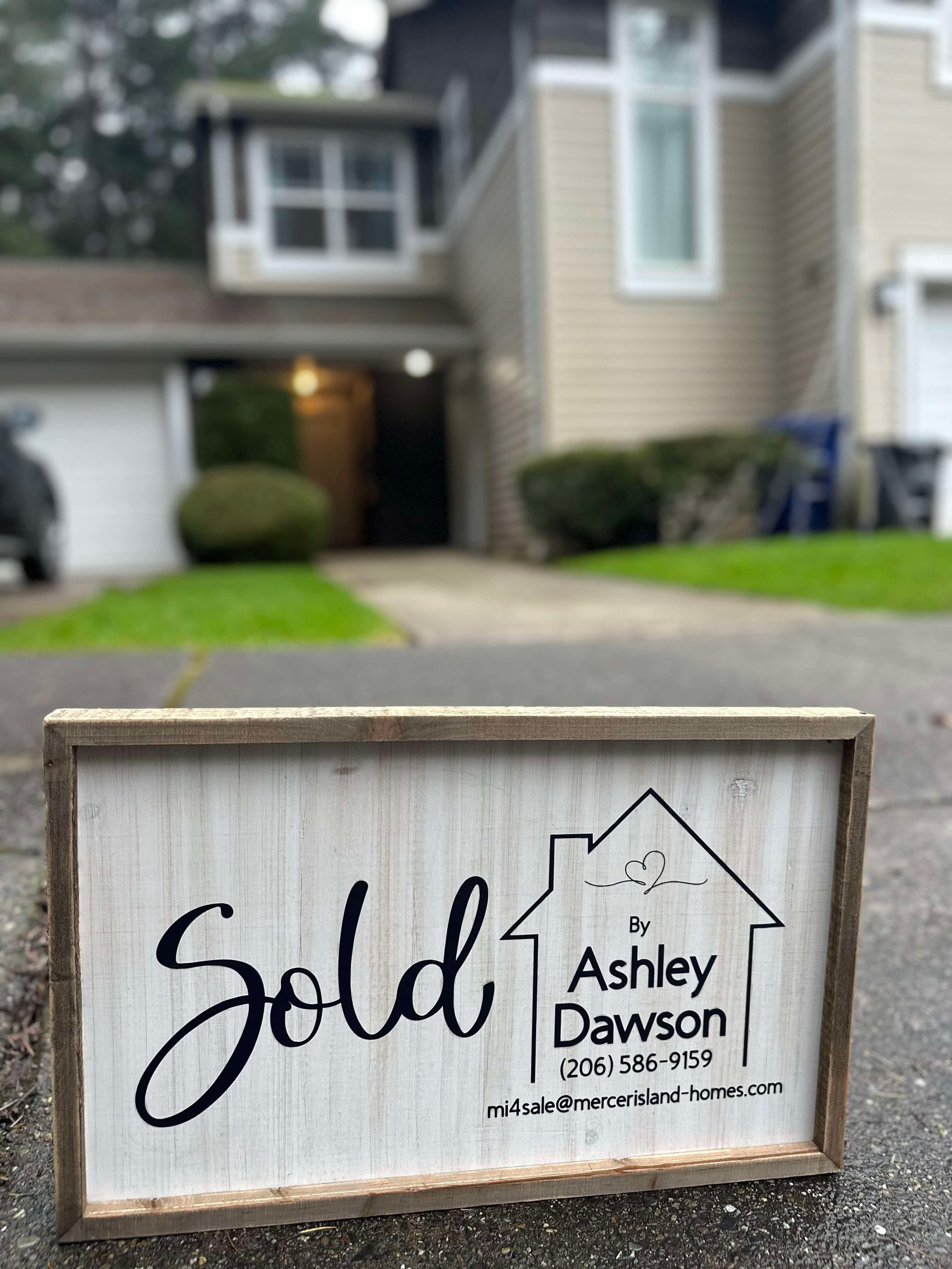 Wooden sign with 'Sold by Ashley Dawson' and contact info in front of a house driveway.