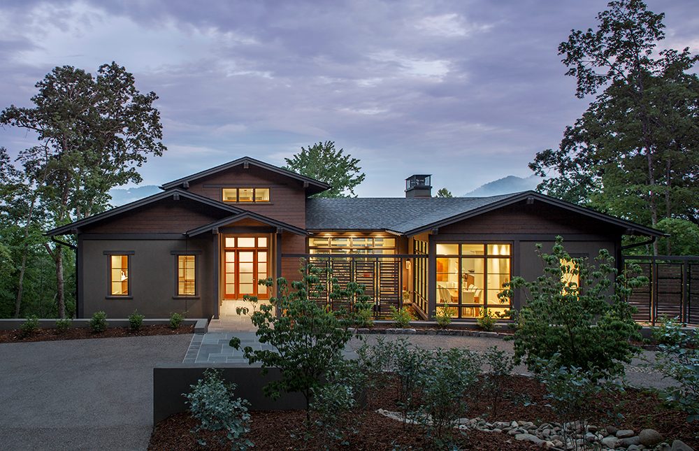 Modern single-story house with large windows and warm lights surrounded by trees at dusk.