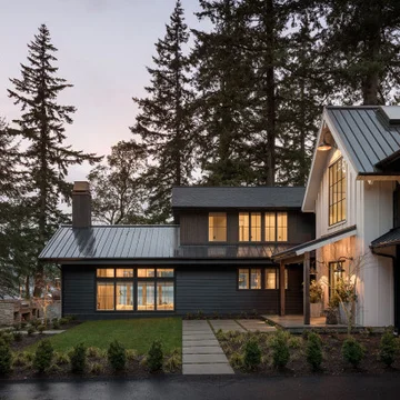 Modern two-story house with dark and white exterior, lit windows, surrounded by tall pine trees at dusk.
